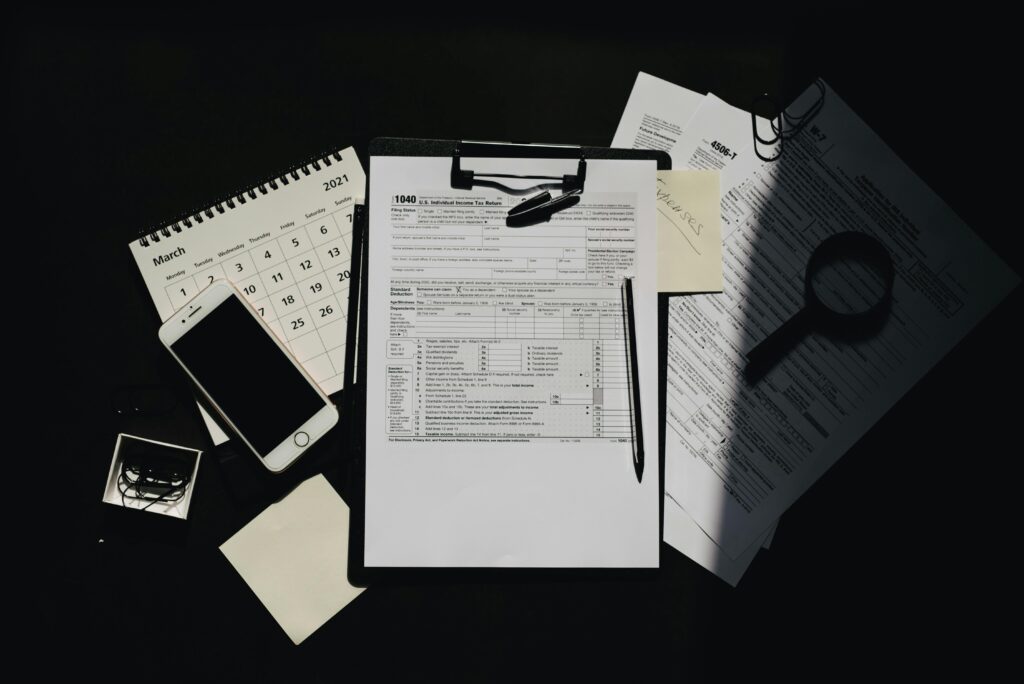 A high-angle shot of a desk with an open calendar, a smartphone, a clipboard with a tax form, a pen, and a magnifying glass. The items are arranged on a dark surface with strong, directional lighting.
